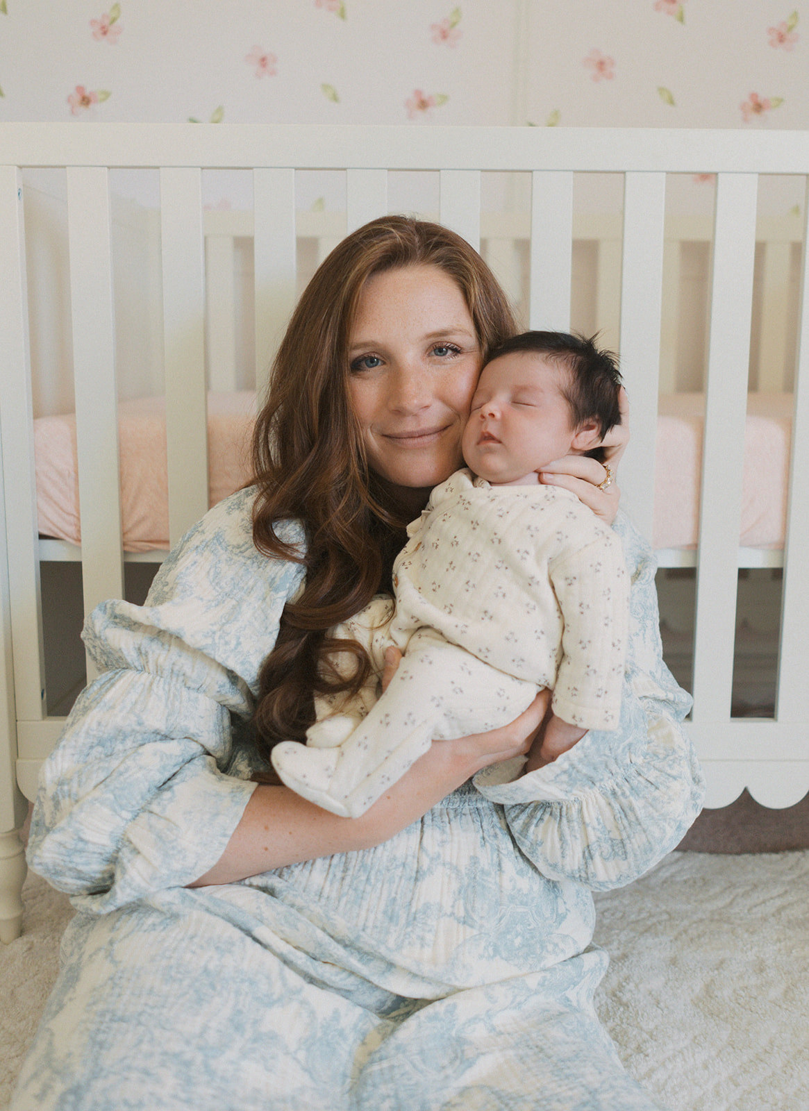 a mother holds her newborn daughter against her cheek as she sits on the floor and smiles at the camera